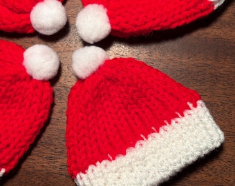May include: Four miniature red and white knit Santa hats with white pom-poms. The hats are made of a chunky knit yarn and are arranged on a dark wood surface. The hats are a festive holiday decoration.