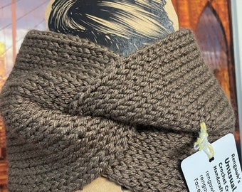 May include: A brown, knitted headband with a twisted design. The headband is handmade and has a tag that reads "Unusual Wedge Crochet Creations". The headband is displayed on a decorative card with drawn eyelashes.