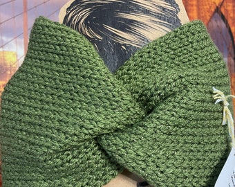 May include: Olive green knitted headband with a twisted knot design. The headband is made of a textured yarn and is displayed on a cardboard backing with a stylized illustration. A small tag is attached.