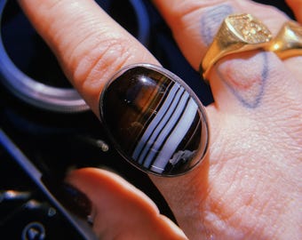 May include: A silver ring featuring a large, oval-shaped stone with brown, white, and black stripes. The ring is worn on a finger, with other gold rings visible in the background. The stone has a polished, glossy finish.