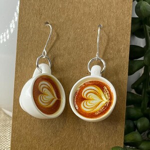 May include: A pair of white coffee cup earrings with silver hooks. Each earring features a detailed latte art design in shades of brown and cream, set against a white cup. The earrings are displayed on a brown card.