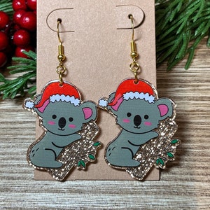 May include: A pair of gold dangle earrings featuring a koala wearing a red Santa hat and holding a gold glitter branch. The koala is gray with pink cheeks and black eyes.