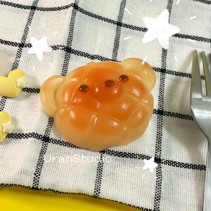 May include: A light orange bear-shaped bread with brown eyes. The bread is on a white and black checkered cloth.