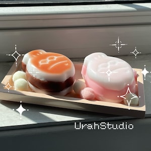 May include: Two decorative soap bars shaped like fish. The fish are on a light brown tray with white decorative balls. The fish are layered with pink, white, and brown soap. The fish are on a white surface.