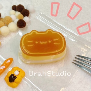 Può includere: Un sacchetto di plastica trasparente contenente un dessert al budino a forma di gatto giallo e marrone. Il budino ha una copertura di caramello marrone e uno strato bianco sottostante. Il dessert è su una superficie bianca con una forchetta e altri piccoli oggetti decorativi.