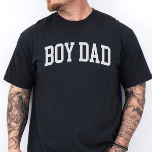 May include: Black t-shirt featuring the white text "BOY DAD" in an arched design. The t-shirt has a crew neck. The person wearing the shirt has tattoos on their arms and is wearing blue jeans.