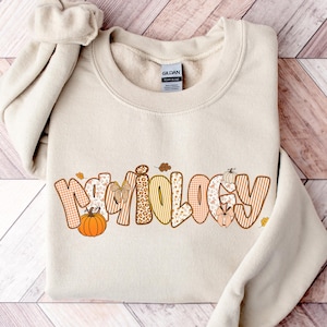 May include: A beige sweatshirt with the word "Radiology" in a fall-themed design. The letters are decorated with pumpkins, leaves, and various patterns in orange, yellow, and brown. The sweatshirt has a crew neck and long sleeves.