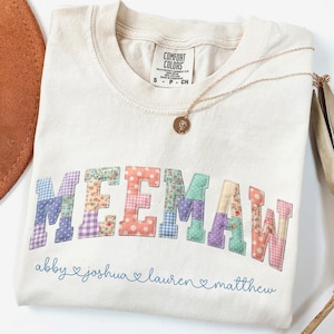 May include: Cream-colored t-shirt with the word "MEEMAW" in patchwork letters. Below the word are the names "abby", "joshua", "lauren", and "matthew". A gold necklace with a pendant rests on the shirt.