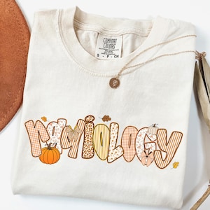 May include: Cream-colored t-shirt with the word "Mythology" in a fall-themed design. The word features pumpkins, leaves, and various patterns. A gold necklace with a pendant is draped on the shirt.