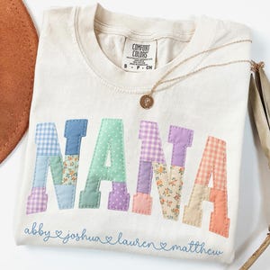 May include: Cream-colored Comfort Colors t-shirt with the word "NANA" appliqued in patchwork letters. The letters are in various pastel colors and patterns. Below the word are the names "abby", "joshua", "lauren", and "matthew". A gold necklace with a pendant rests on the shirt.
