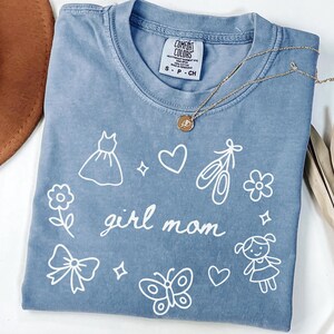 May include: A blue Comfort Colors t-shirt with the words "girl mom" and various white illustrations, including a dress, hearts, ballet shoes, and a butterfly. A gold necklace with a pendant rests on the shirt.