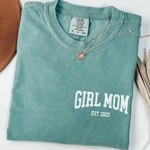 Op de afbeelding: Een teal Comfort Colors T-shirt met de tekst "GIRL MOM EST. 2025" in het wit. Een goudkleurige ketting met een hanger is over het shirt gedrapeerd. Een bruine hoed en gedroogde plantenstelen staan op de achtergrond.