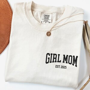 Op de afbeelding: Cr&egrave;mekleurig T-shirt met de tekst "GIRL MOM EST. 2025" in het zwart gedrukt. Een gouden ketting met een hanger ligt op het shirt. Een bruine hoed en gedroogde plantenstengels zijn op de achtergrond.