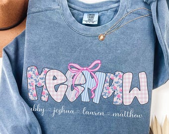 May include: A blue sweatshirt with the word "MEOW" in a playful font, decorated with a pink bow and floral and striped patterns. The names "abby joshua lauren matthew" are printed below the word.