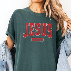 Pu&ograve; includere: Una maglietta verde scuro con la parola "JESUS" in lettere rosse a blocchi e il testo "HE IS RISEN" in un rettangolo rosso pi&ugrave; piccolo sotto.
