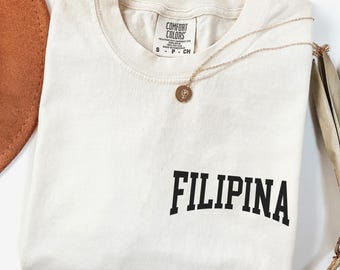 May include: Cream-colored t-shirt with the word "FILIPINA" printed in bold black letters. A gold necklace with a pendant rests on the shirt. A brown hat and dried plant are in the background.