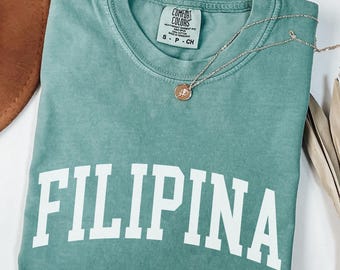 May include: A seafoam green t-shirt with the word "FILIPINA" in bold white letters. The shirt has a crew neck and a small tag that reads "COMFORT COLORS". A silver necklace with a pendant is draped across the shirt.