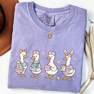 May include: A lilac Comfort Colors t-shirt featuring a graphic of four cartoon ducks dressed as healthcare workers. One duck holds a basket of eggs, another has a stethoscope, a third holds a sign that says "NURSE LIFE", and the last wears bunny ears.