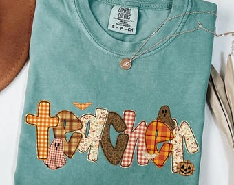 May include: A teal Comfort Colors t-shirt with the word "teacher" appliqued in various fall-themed patterns, including plaid and floral designs. The shirt features a small ghost and pumpkin graphic. A silver necklace with a pendant is also visible.