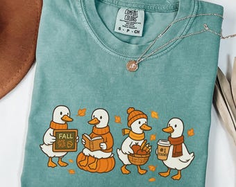 May include: A teal Comfort Colors t-shirt featuring a fall-themed graphic of four ducks. The ducks are wearing scarves and hats, holding a sign that says "FALL", reading a book, carrying a basket of apples, and holding a coffee cup that says "#1 Teacher".