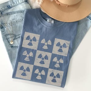 May include: A blue t-shirt with a white checkered pattern featuring a repeated image of a radiation symbol. The shirt is folded and laid on a pair of blue jeans and a brown hat.