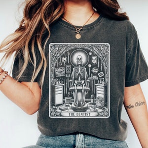 May include: A dark gray t-shirt with a graphic print of a skeleton sitting in a dentist's chair surrounded by dental tools. The graphic is in black and white and has the text "The Dentist" at the bottom.
