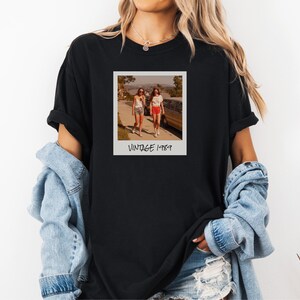 Pode incluir: T-shirt preto com uma fotografia em estilo vintage de duas pessoas a caminhar na rua. A fotografia est&aacute; numa moldura branca com o texto "VINTAGE 1989" por baixo. A pessoa que usa a t-shirt tem cabelo loiro comprido.