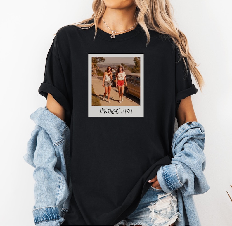 Pode incluir: T-shirt preto com uma fotografia em estilo vintage de duas pessoas a caminhar na rua. A fotografia est&aacute; numa moldura branca com o texto "VINTAGE 1989" por baixo. A pessoa que usa a t-shirt tem cabelo loiro comprido.