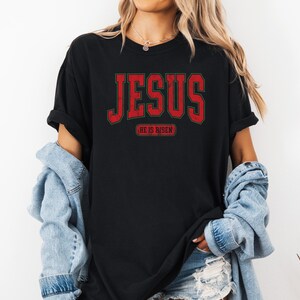Pu&ograve; includere: Maglietta nera con una grafica rossa "JESUS" e il testo "HE IS RISEN" in un carattere pi&ugrave; piccolo sotto.
