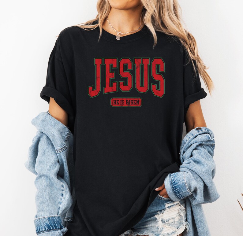 Pu&ograve; includere: Maglietta nera con una grafica rossa "JESUS" e il testo "HE IS RISEN" in un carattere pi&ugrave; piccolo sotto.