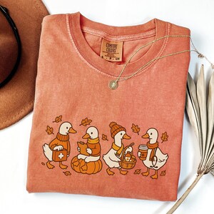 May include: A coral Comfort Colors t-shirt featuring a whimsical illustration of four ducks dressed in fall attire. One duck carries a first aid kit, another reads a book, and others hold a basket and coffee. A gold necklace and a brown hat are also visible.
