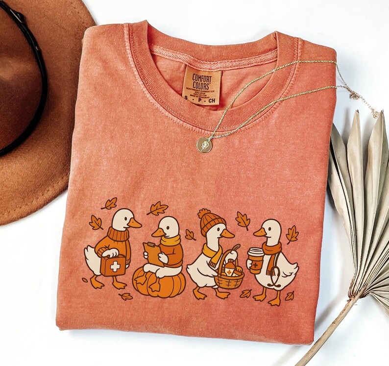 May include: A coral Comfort Colors t-shirt featuring a whimsical illustration of four ducks dressed in fall attire. One duck carries a first aid kit, another reads a book, and others hold a basket and coffee. A gold necklace and a brown hat are also visible.
