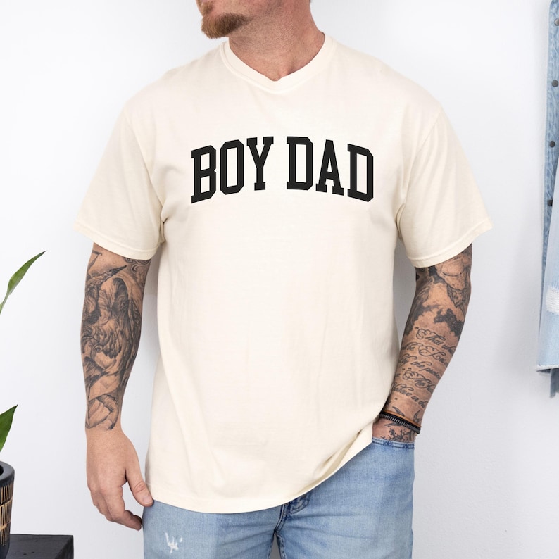 May include: Cream-coloured v-neck t-shirt with the text "BOY DAD" in bold, black letters across the chest. The shirt has short sleeves and a relaxed fit. The person is wearing blue jeans.