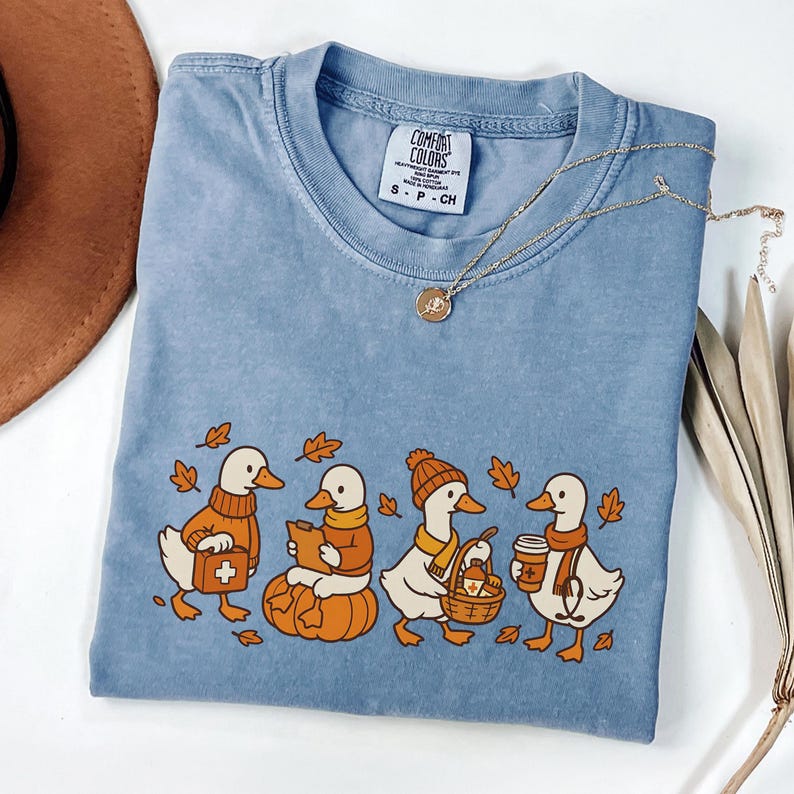 May include: A light blue Comfort Colors t-shirt featuring a fall-themed graphic of four ducks. The ducks are dressed in autumn attire, carrying items like a first aid kit, a book, a basket, and a coffee cup. A gold necklace and a brown hat are also visible.