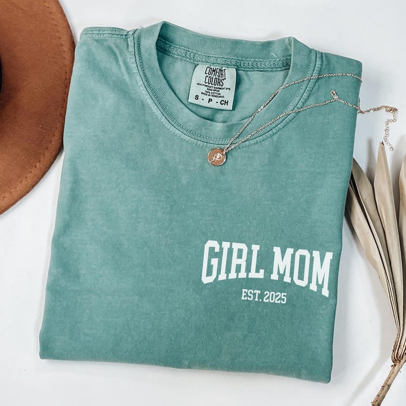 Op de afbeelding: Een teal Comfort Colors T-shirt met de tekst "GIRL MOM EST. 2025" in het wit. Een goudkleurige ketting met een hanger is over het shirt gedrapeerd. Een bruine hoed en gedroogde plantenstelen staan op de achtergrond.