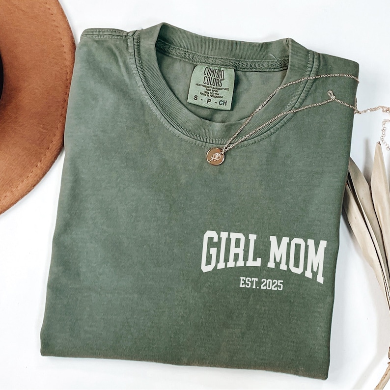 Op de afbeelding: Een olijfgroen t-shirt met de witte tekst "GIRL MOM EST. 2025". Een gouden ketting met een hanger ligt over het shirt. Het shirt is op een wit oppervlak gevouwen.
