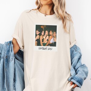 Pode incluir: Camiseta creme com estampa fotogr&aacute;fica em estilo vintage. A foto mostra cinco pessoas e o texto "VINTAGE 2021". A camiseta tem mangas compridas e um corte folgado. Uma jaqueta jeans &eacute; usada.