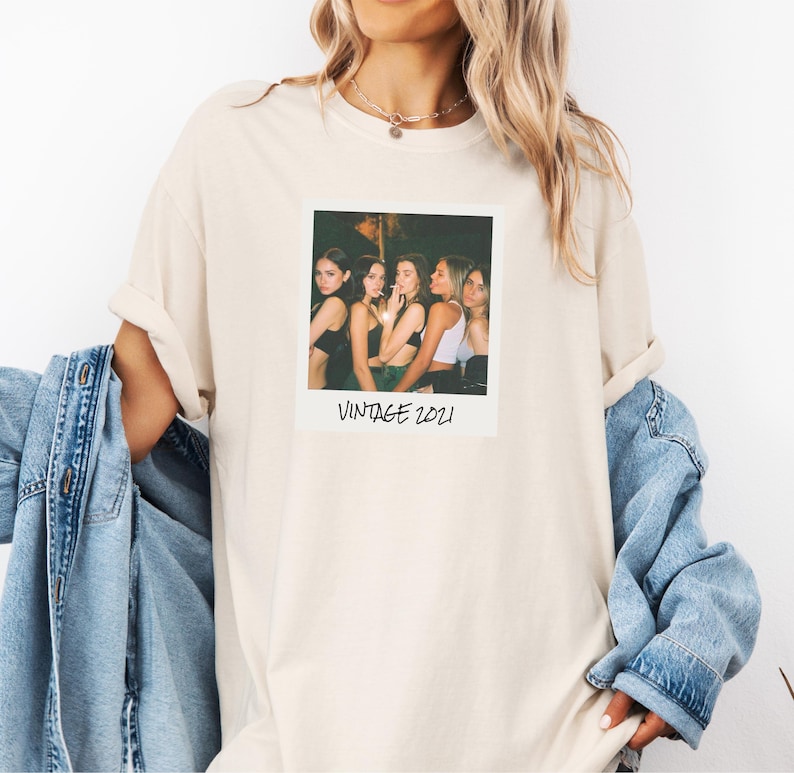 Pode incluir: Camiseta creme com estampa fotogr&aacute;fica em estilo vintage. A foto mostra cinco pessoas e o texto "VINTAGE 2021". A camiseta tem mangas compridas e um corte folgado. Uma jaqueta jeans &eacute; usada.