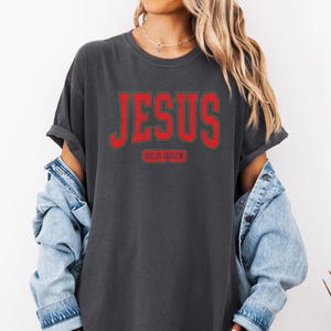 Pu&ograve; includere: Una maglietta grigia scura con una grafica rossa "JESUS" e il testo "HE IS RISEN" in un carattere pi&ugrave; piccolo sotto.