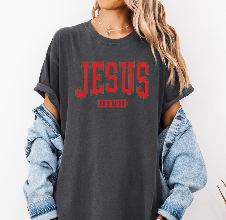 Pu&ograve; includere: Una maglietta grigia scura con una grafica rossa "JESUS" e il testo "HE IS RISEN" in un carattere pi&ugrave; piccolo sotto.