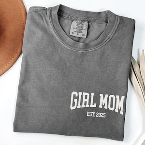 Op de afbeelding: Een grijs Comfort Colors T-shirt met de woorden "GIRL MOM EST. 2025" in het wit gedrukt. Het shirt is opgevouwen en heeft een label bij de halslijn. Een bruine hoed en decoratieve bladeren zijn gedeeltelijk zichtbaar op de achtergrond.