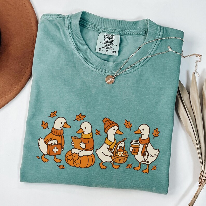 May include: Teal Comfort Colors t-shirt featuring a fall-themed graphic of ducks dressed in autumn attire. The ducks are wearing sweaters, scarves, and hats, and are surrounded by fall leaves. A silver necklace with a pendant is also visible.