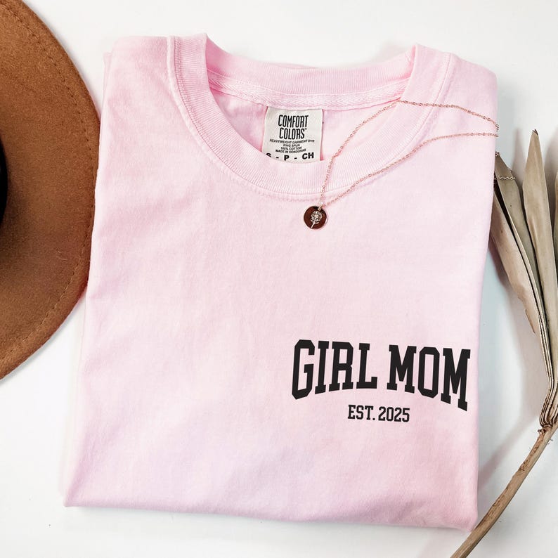 Op de afbeelding: Een lichtroze t-shirt met de tekst "GIRL MOM EST. 2025" in het zwart gedrukt. Een gouden ketting met een hanger ligt over het shirt. Een bruine hoed en gedroogde planten staan op de achtergrond.