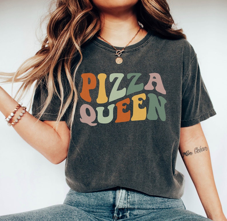 May include: A dark gray t-shirt with the text "PIZZA QUEEN" in colorful retro-style lettering.