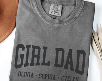 May include: A gray Comfort Colors t-shirt with the words "GIRL DAD" in bold, black letters. Below the text are the names "OLIVIA - SOPHIA - EVELYN". The shirt is folded and lies on a white surface. A brown hat is visible in the upper left corner.