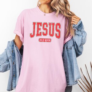 Pu&ograve; includere: Maglietta rosa con una grafica rossa "JESUS" in stile vintage universitario. Il testo "HE IS RISEN" &egrave; stampato sotto la grafica.