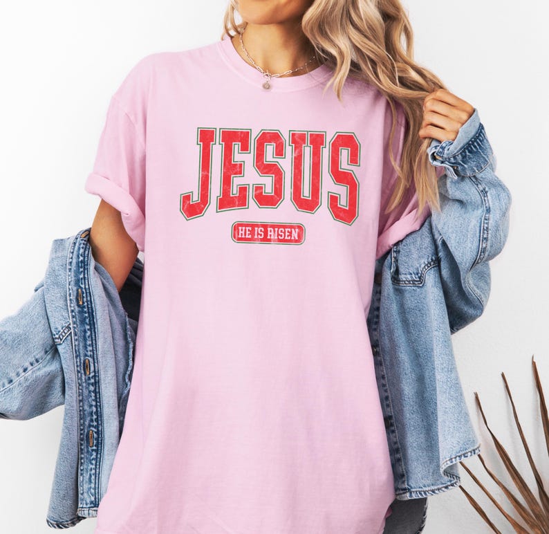 Pu&ograve; includere: Maglietta rosa con una grafica rossa "JESUS" in stile vintage universitario. Il testo "HE IS RISEN" &egrave; stampato sotto la grafica.