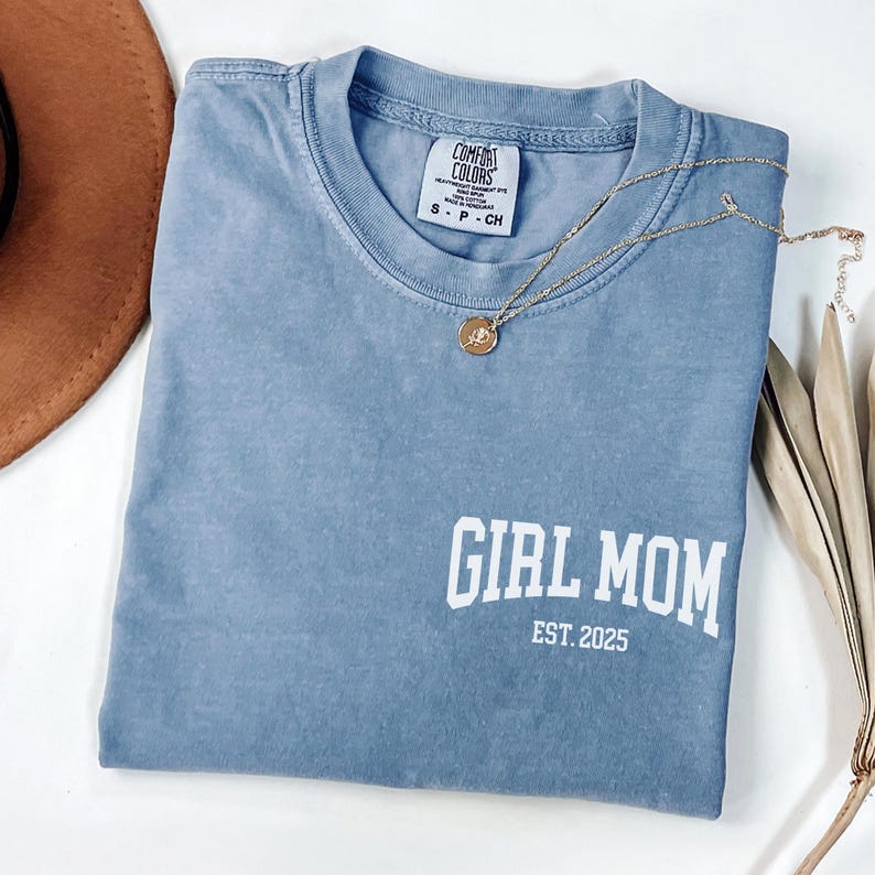 Op de afbeelding: Een blauw Comfort Colors T-shirt met de tekst "GIRL MOM EST. 2025" in het wit. Een gouden ketting met een hanger ligt op het shirt. Een bruine hoed en gedroogde plantenstelen staan op de achtergrond.