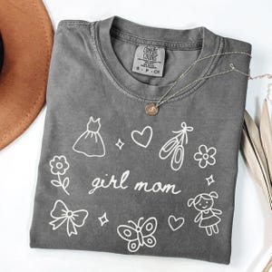 May include: A gray Comfort Colors t-shirt with the words "girl mom" in script, surrounded by white line drawings of a dress, ballet shoes, flowers, a bow, a butterfly, and a girl. A silver necklace with a pendant is draped across the shirt.