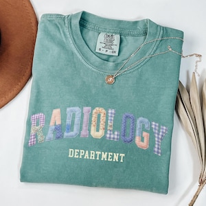 May include: A teal green t-shirt with the word "RADIOLOGY" in patchwork letters and "DEPARTMENT" printed below. A silver necklace with a pendant rests on the shirt. The shirt is folded and placed on a white surface.